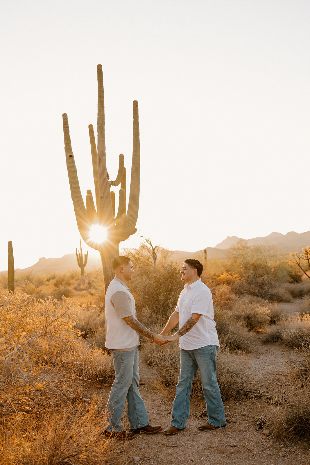 Two men holding hands beneath a saguaro in the desert, the warm light casting long shadows.