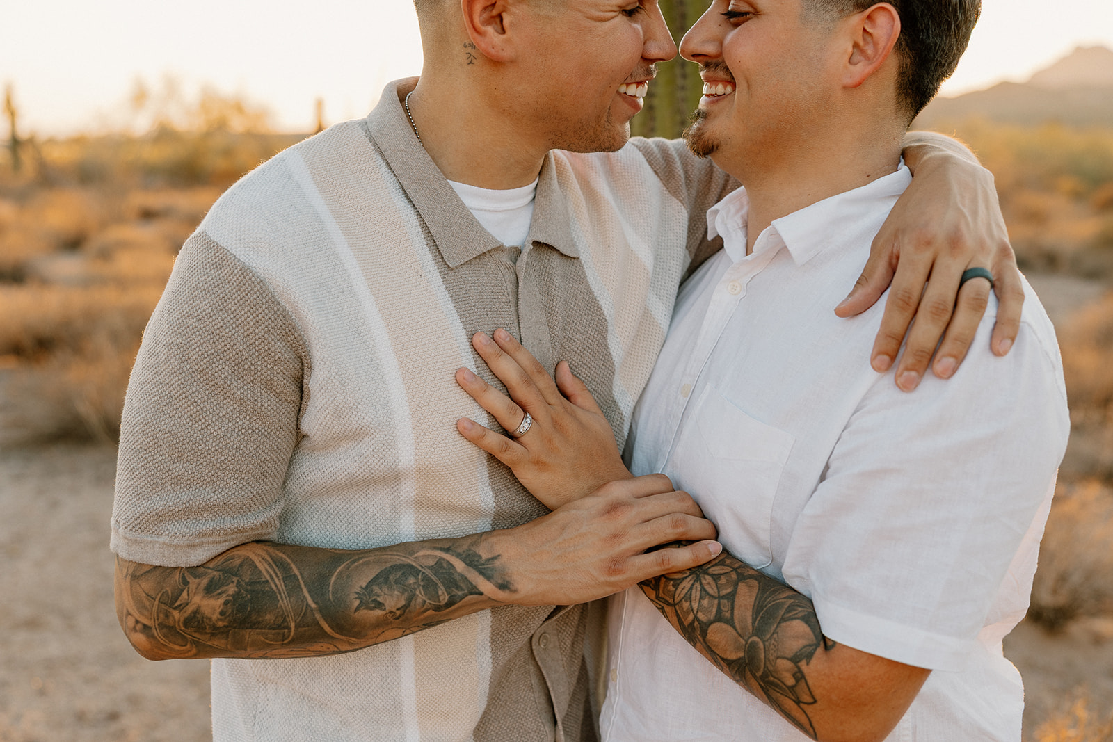 Two grooms laughing and embracing during their desert engagement photos, showcasing tattoos and matching smiles.