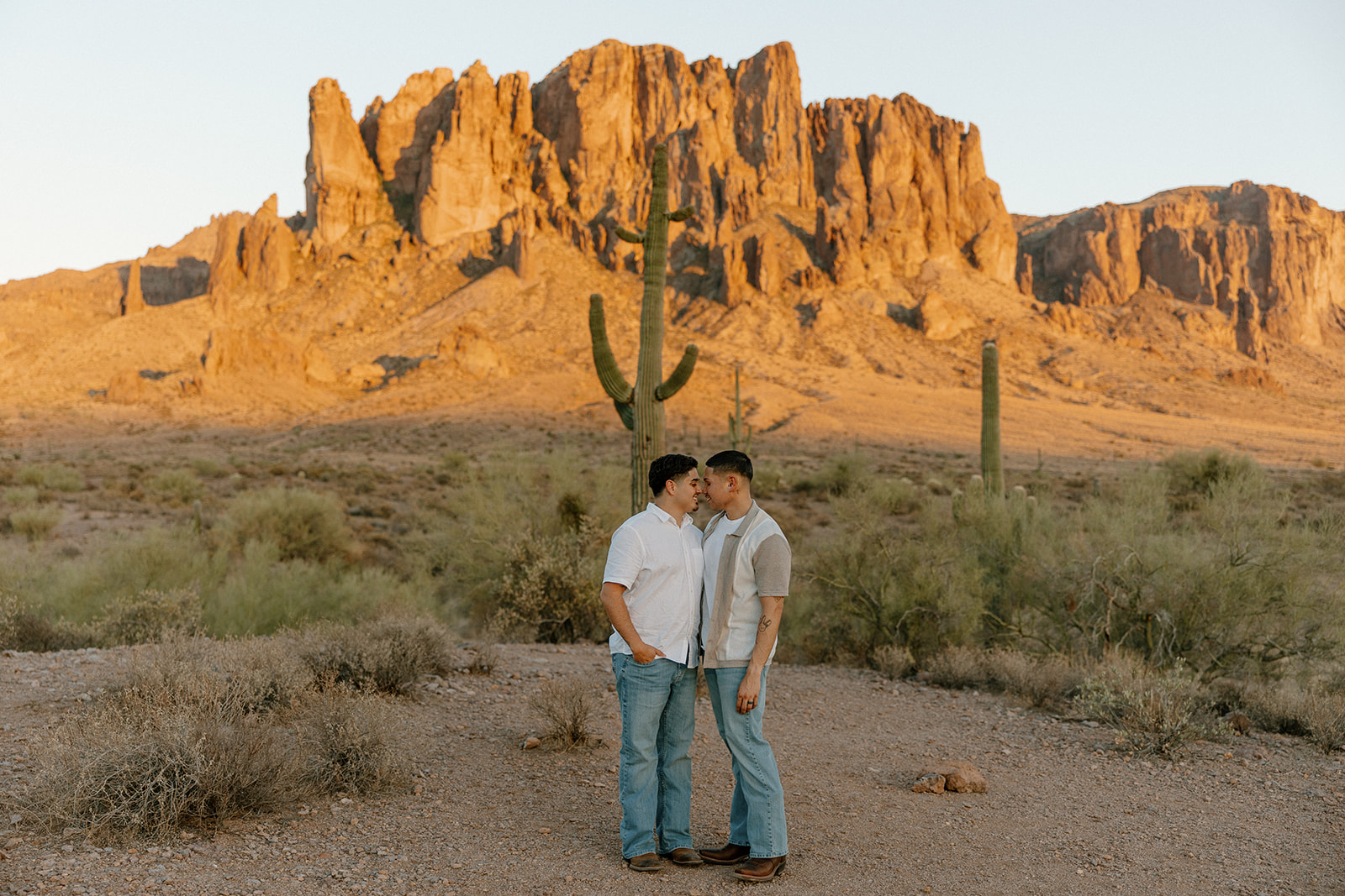 Two grooms standing forehead-to-forehead in front of golden cliffs and towering cacti in the Arizona desert.