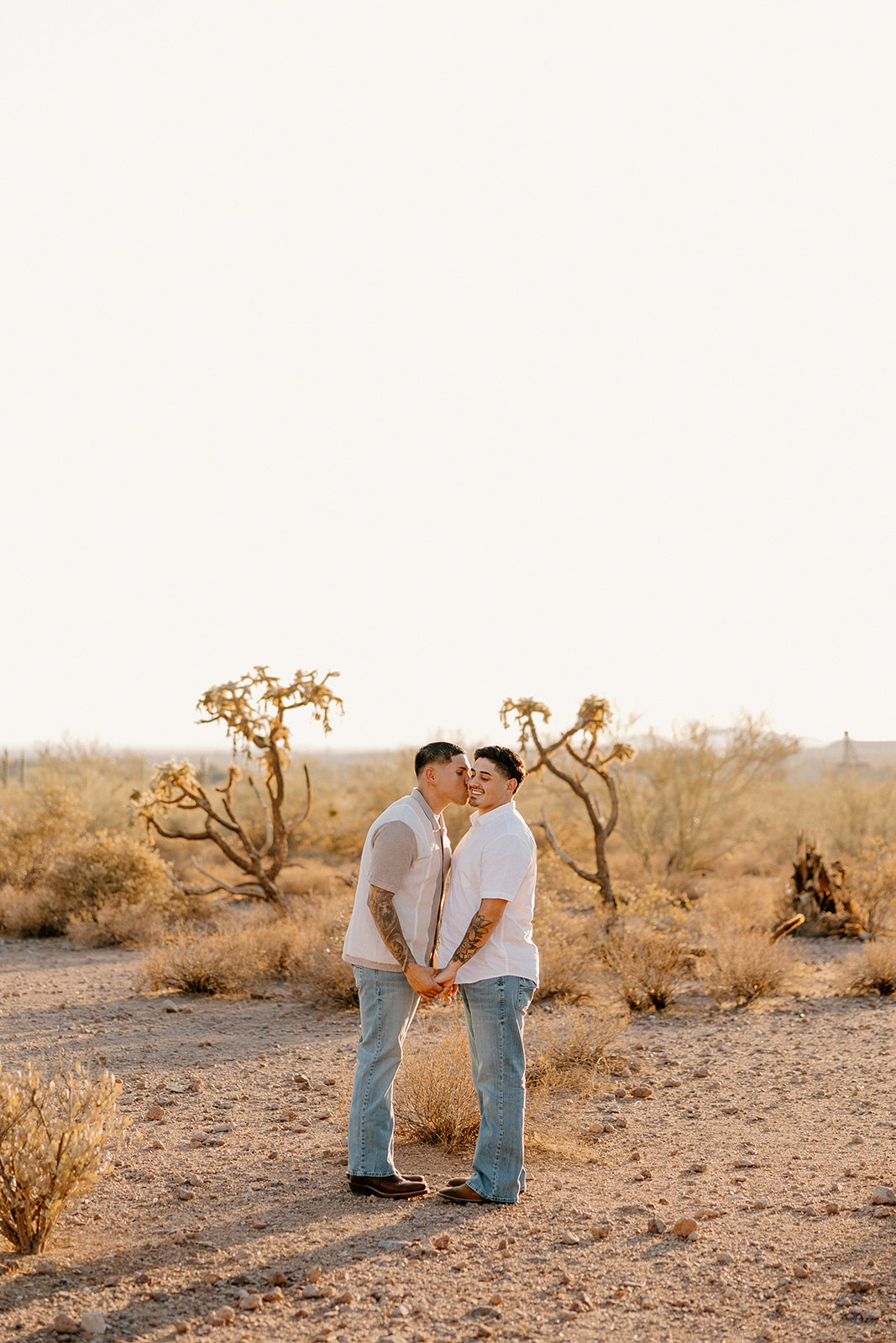Playful and warm desert engagement photo of two men kissing while holding hands in the middle of sun-drenched Arizona terrain.