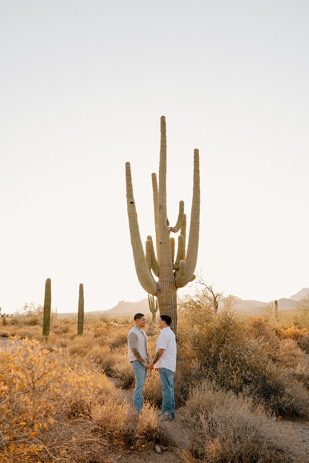 Sun peeking through a saguaro as a couple holds hands during golden-hour desert engagement photos.