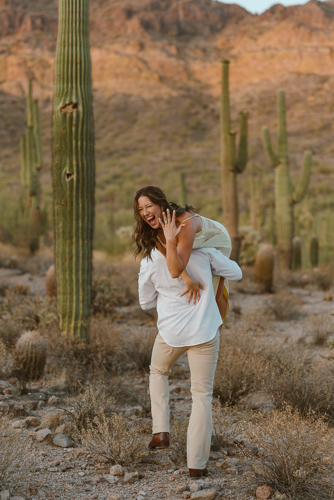 Man lifting his fiancée playfully through the desert, her joy radiating as she shows off her engagement ring among the cacti.