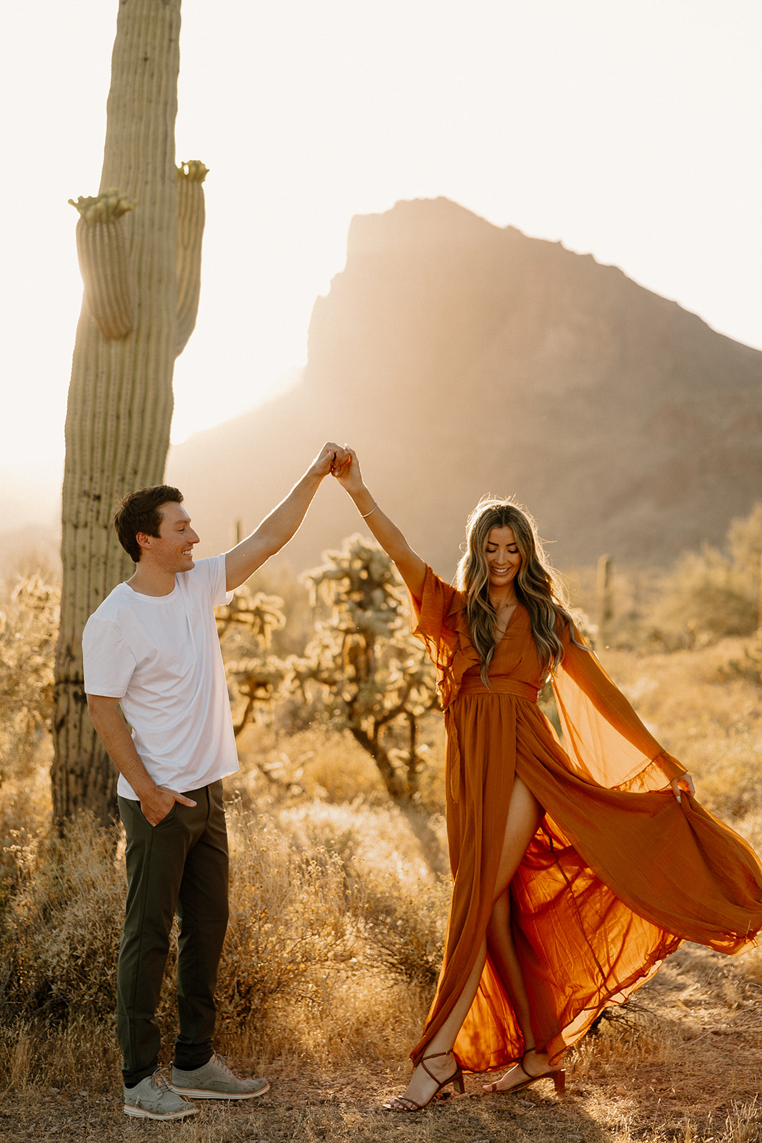 Golden-hour desert engagement photo of a woman in a flowing rust-colored dress and wide-brim hat, looking over her shoulder as her partner faces the mountains.