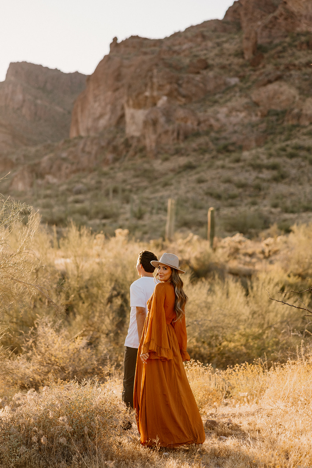 Sunlit desert engagement photo of a couple dancing between towering cacti, her orange dress billowing with the movement.