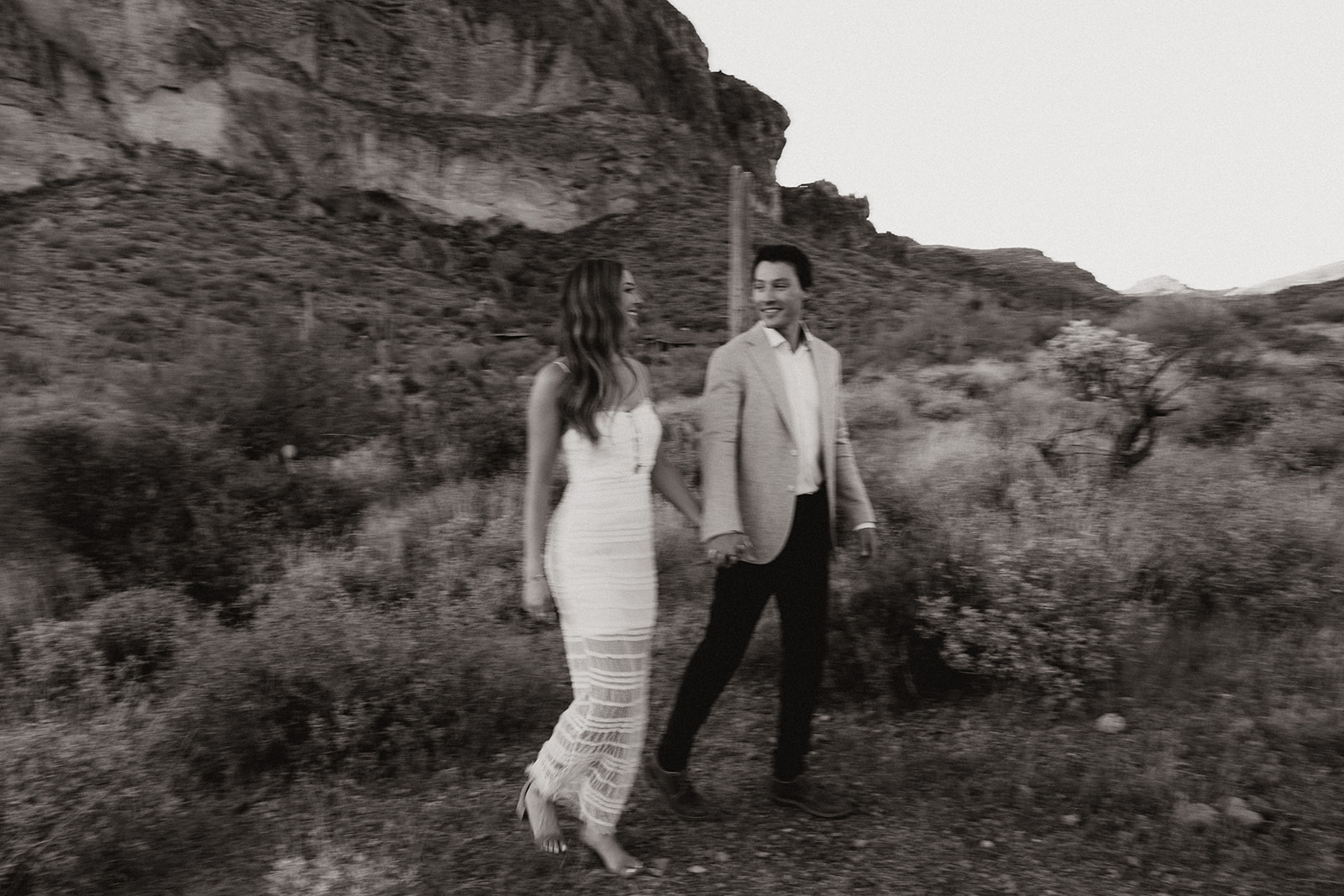 Romantic black-and-white motion blur of a couple walking hand-in-hand through a desert landscape in formal attire.