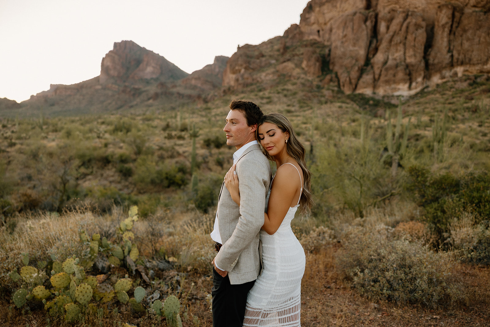 Romantic desert engagement photo of a woman in a white lace dress hugging her partner with mountain views in the background.
