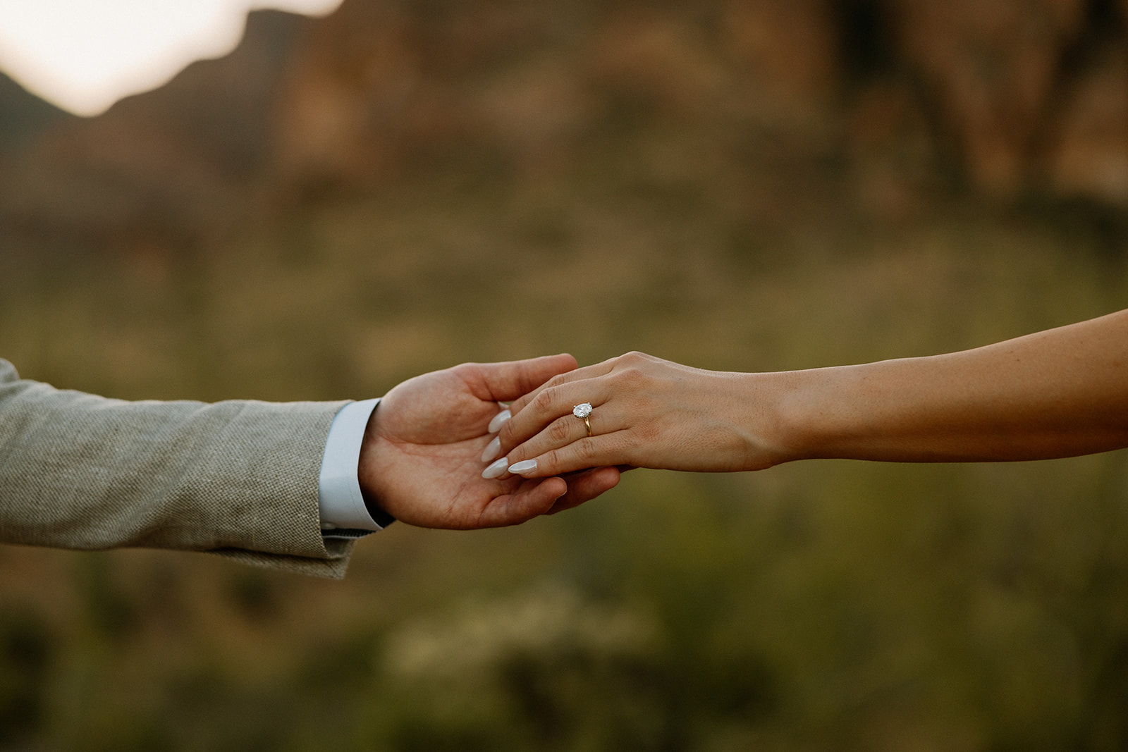 Close-up of hands reaching out, highlighting a sparkling engagement ring against a blurred desert mountain backdrop.