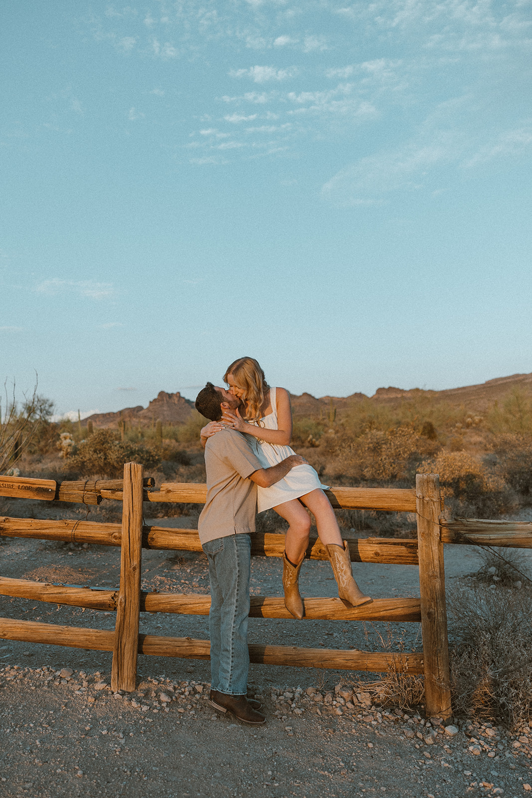 Couple sharing a playful kiss as she sits on a wooden fence in the desert during golden hour.