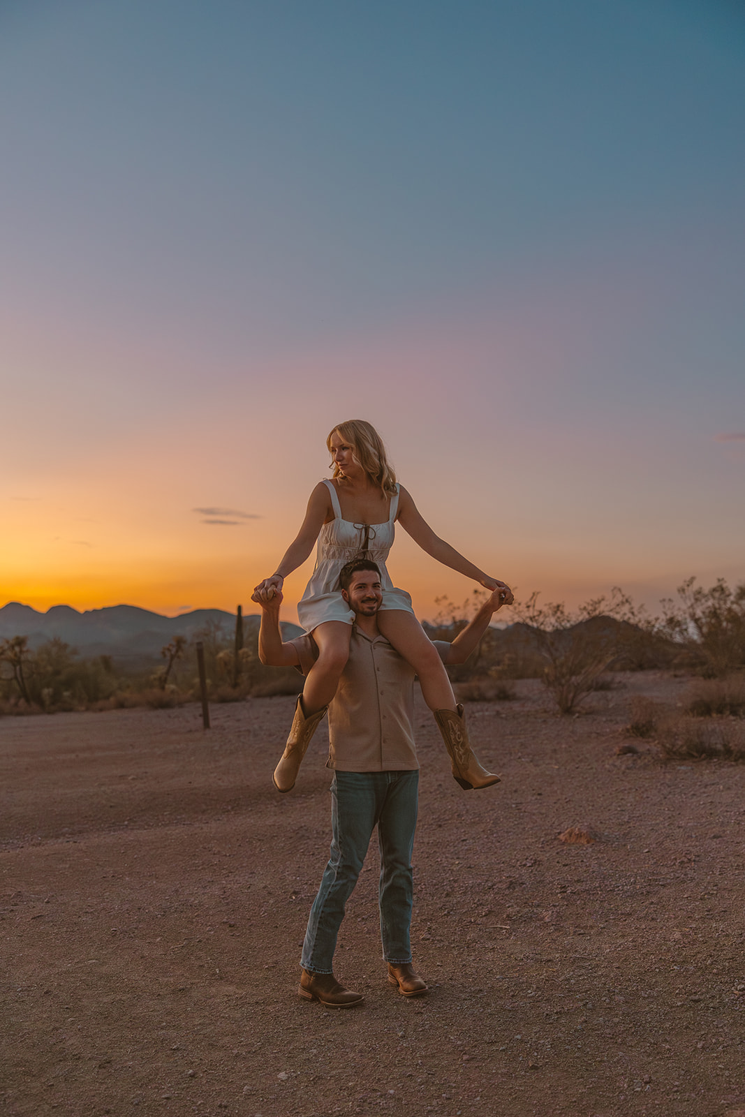 A couple holding hands and laughing during their desert engagement photos, framed by pastel skies and rugged Arizona terrain.