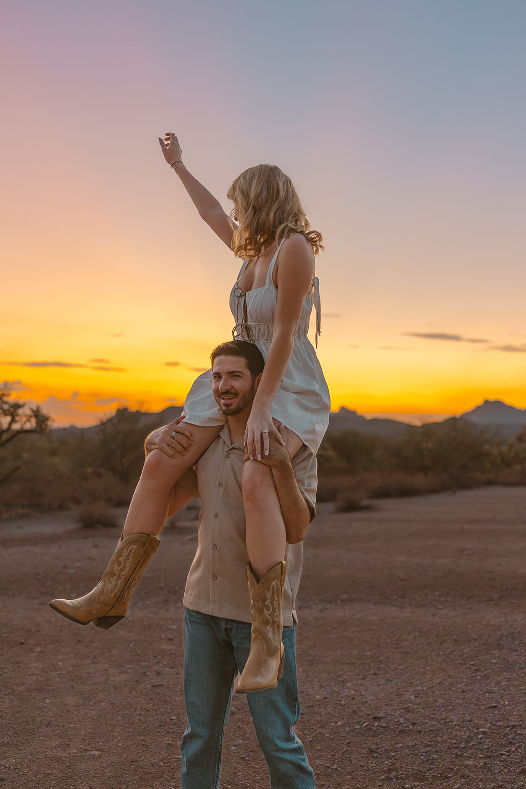 Playful moment during desert engagement photos, with a woman in a white dress and boots sitting on her partner’s shoulders at sunset.