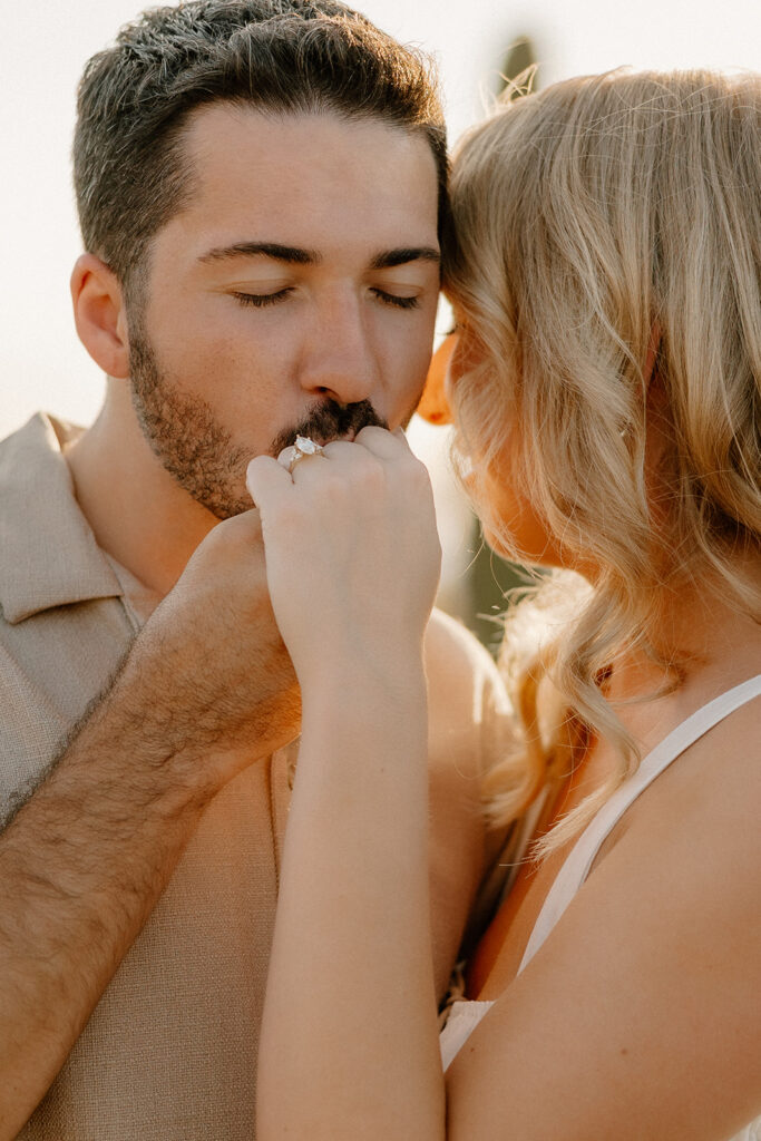 Man gently kissing his partner’s hand, showing off her engagement ring in soft golden light.