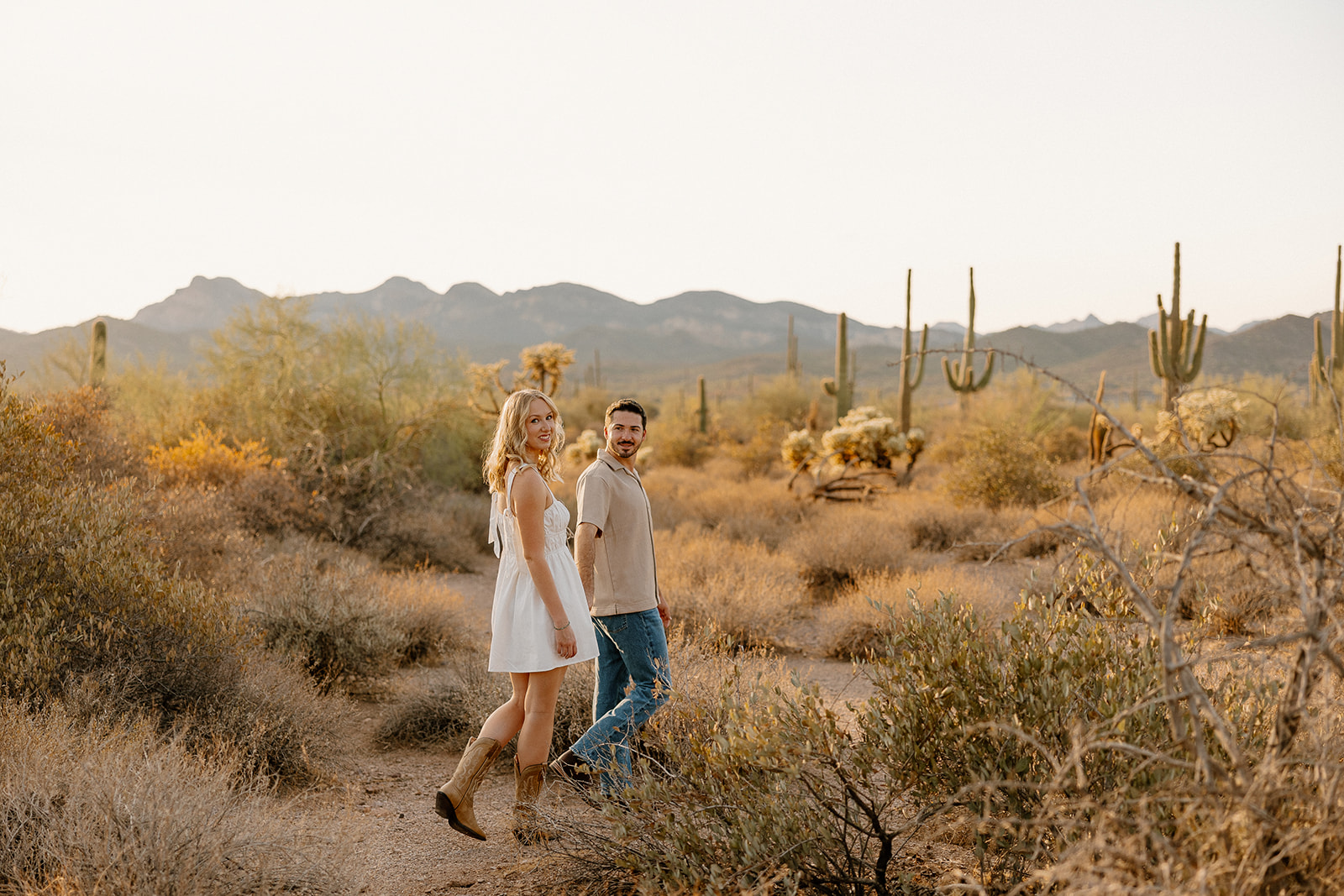 Smiling couple walking through a cactus-dotted trail at sunset during their desert engagement photos.