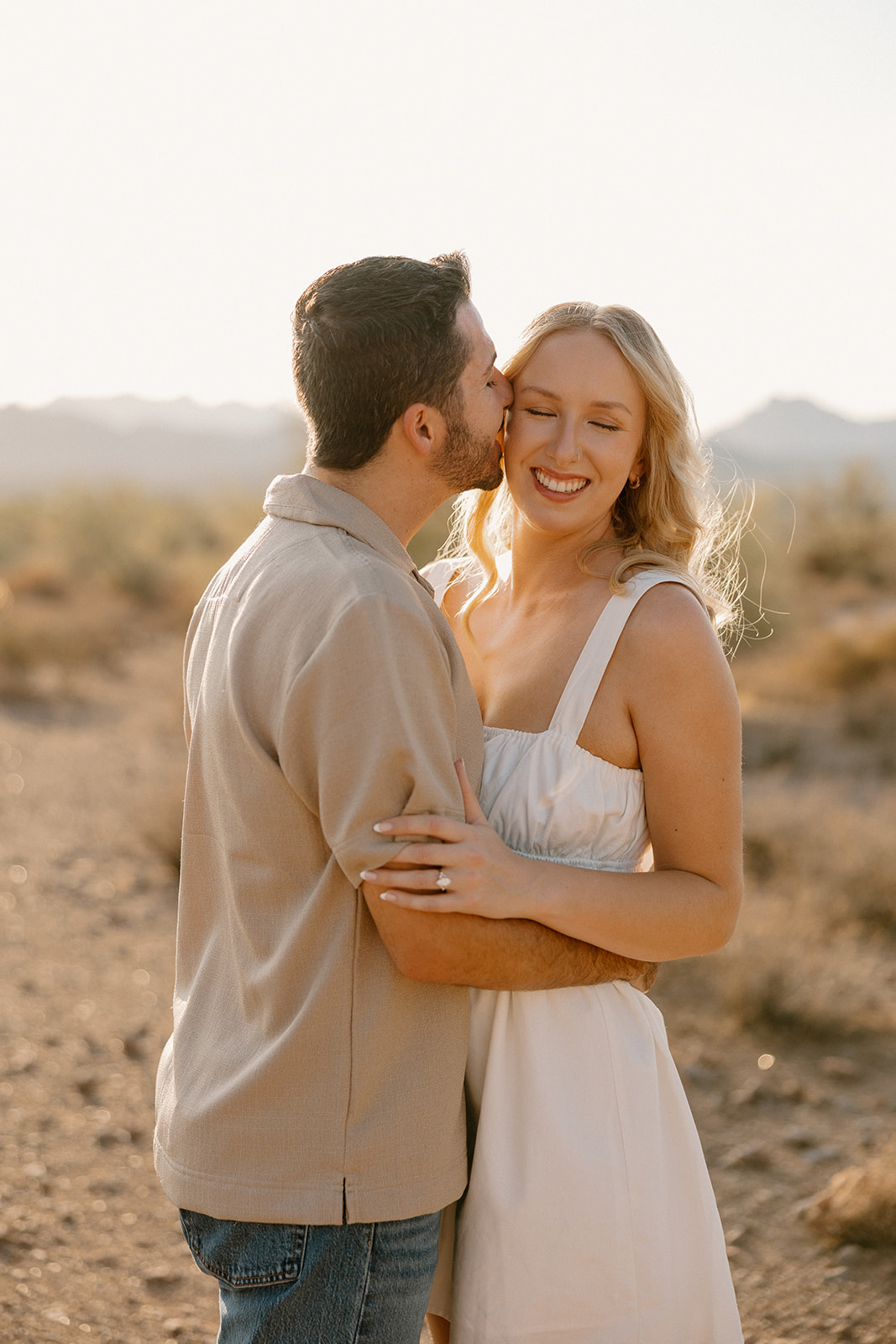 Man kissing his fiancée on the cheek as she smiles, surrounded by soft light and desert haze.