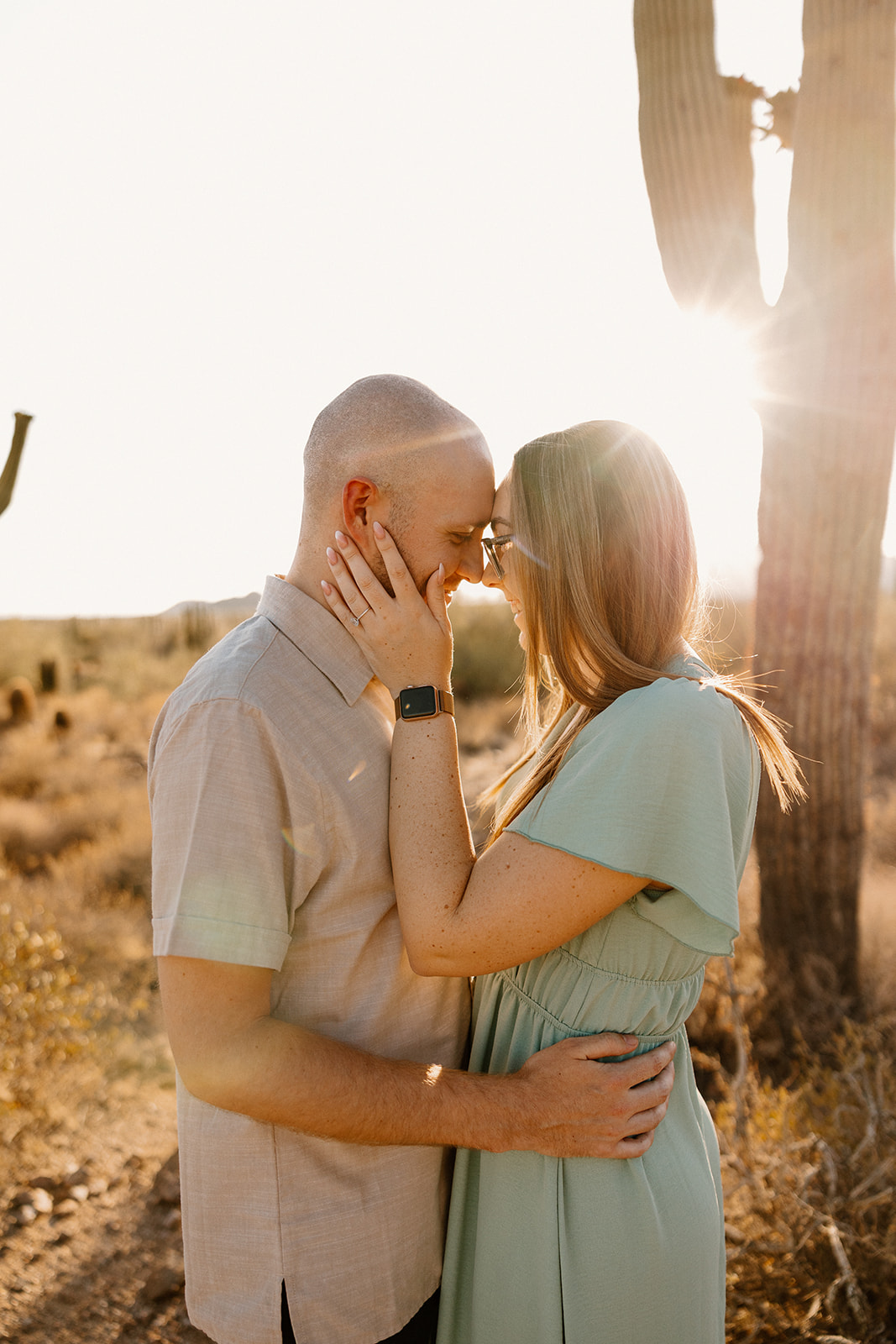 Tender moment during desert engagement photos, as a couple leans in forehead to forehead with sunlight flaring through a cactus.