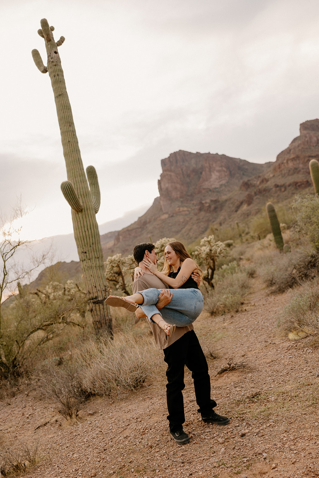Man lifting his fiancée off the ground with both of them laughing in the middle of their desert engagement photos at the base of towering cacti.