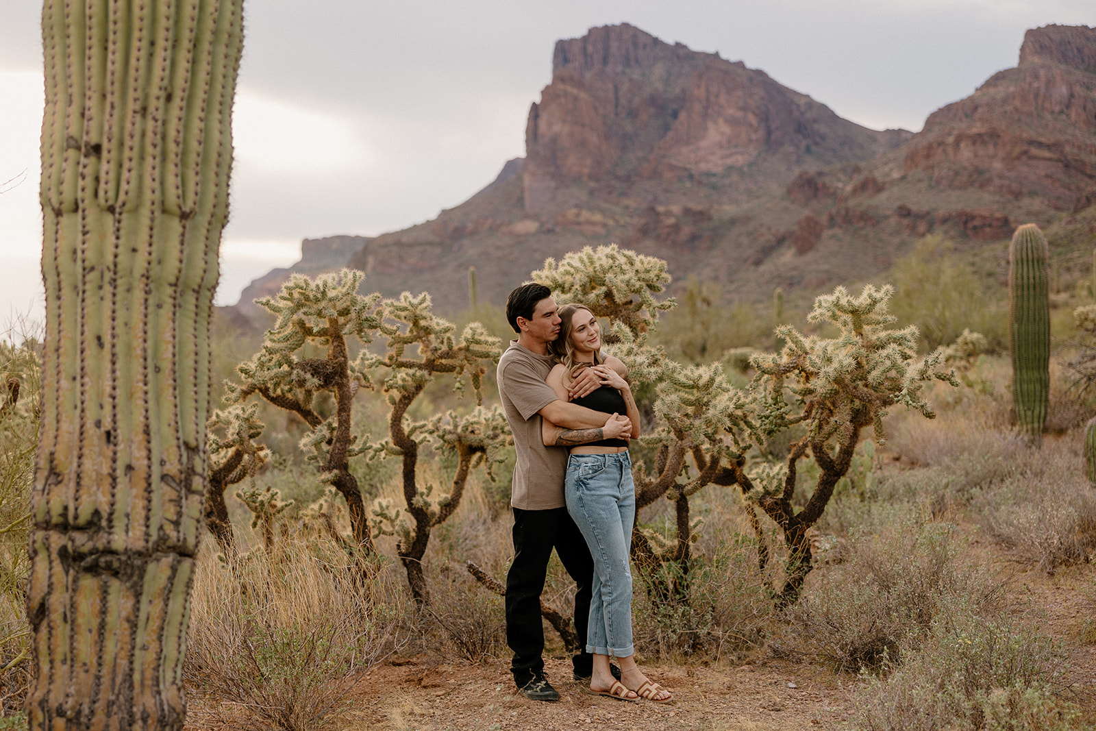 A couple wrapped in each other’s arms during their desert engagement photos at the base of rugged Superstition Mountains.
