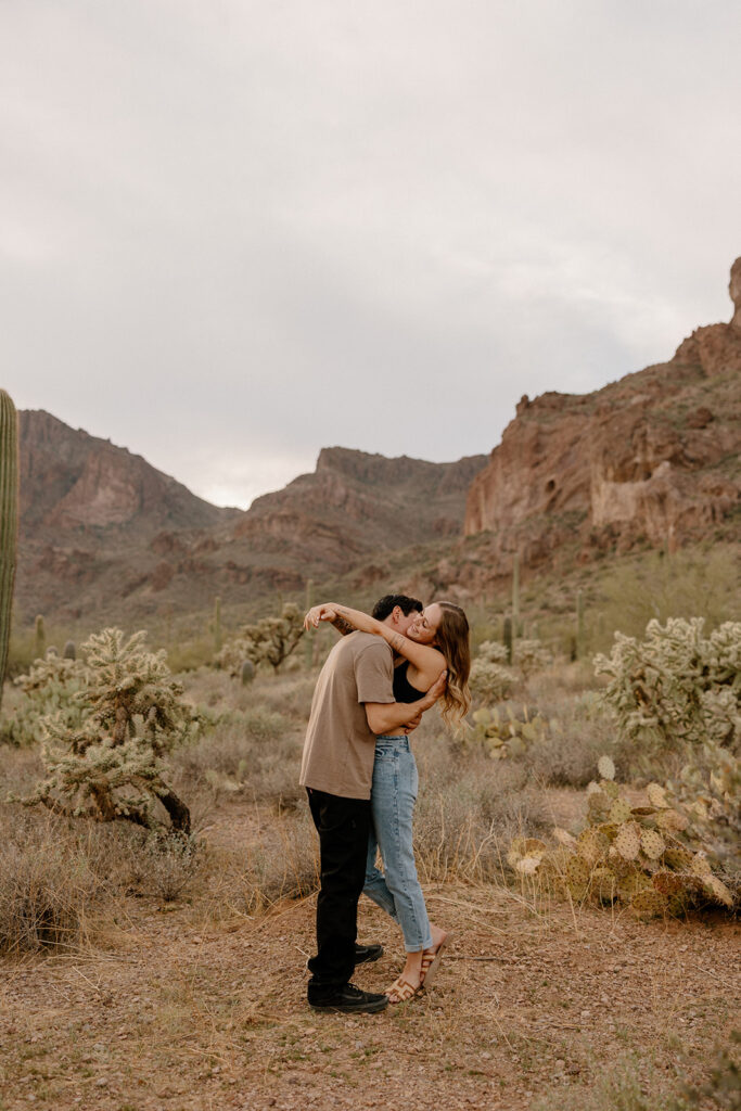 Candid kiss surrounded by desert cactus and rugged mountain peaks during desert engagement photos.