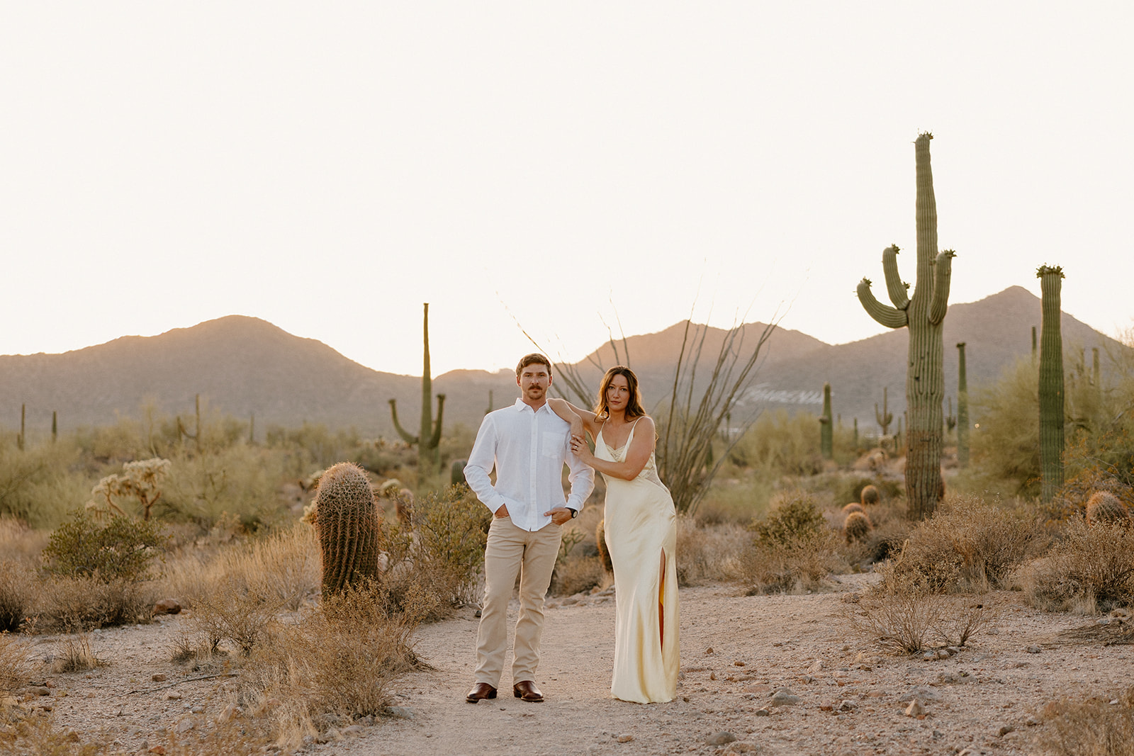 Editorial-style desert engagement photo of a stylish couple standing confidently among saguaros at dusk.