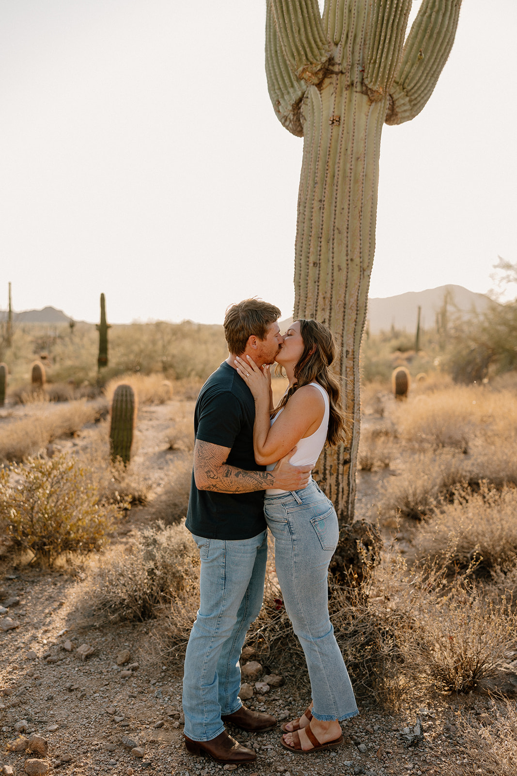 Couple sharing a kiss under a tall saguaro during golden hour in their desert engagement photos.