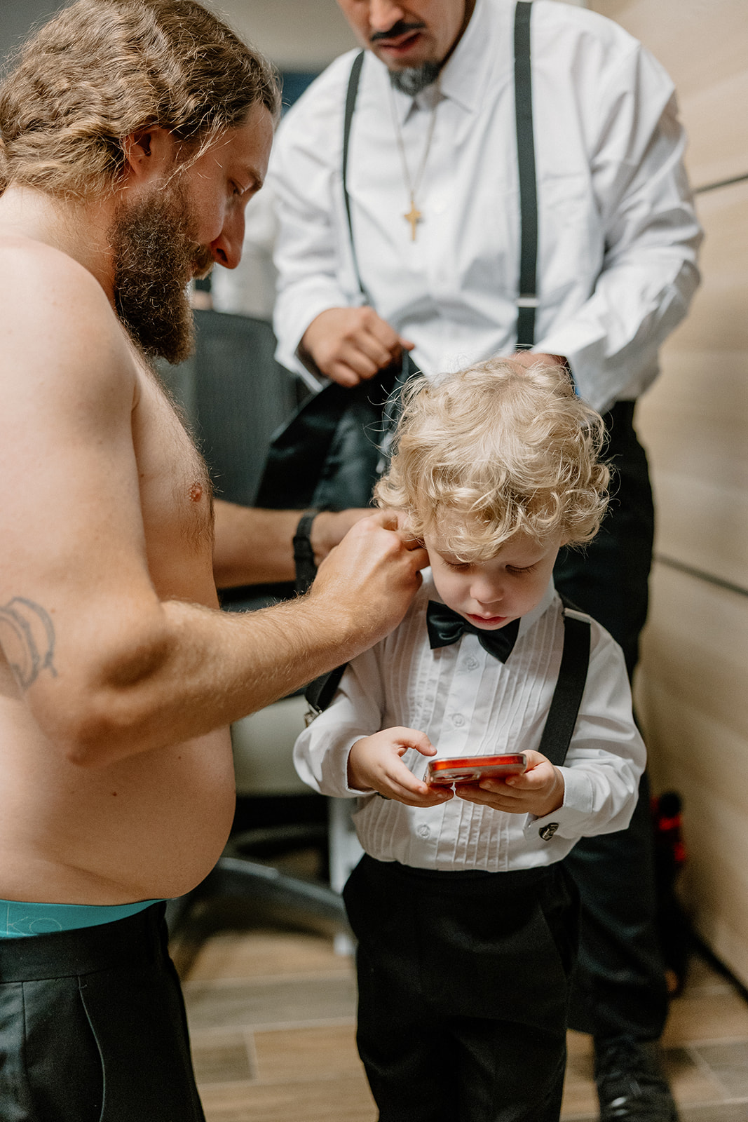 The ring bearer watches a phone intently while the groom adjusts his collar during the getting ready chaos at an Arizona wedding venue.