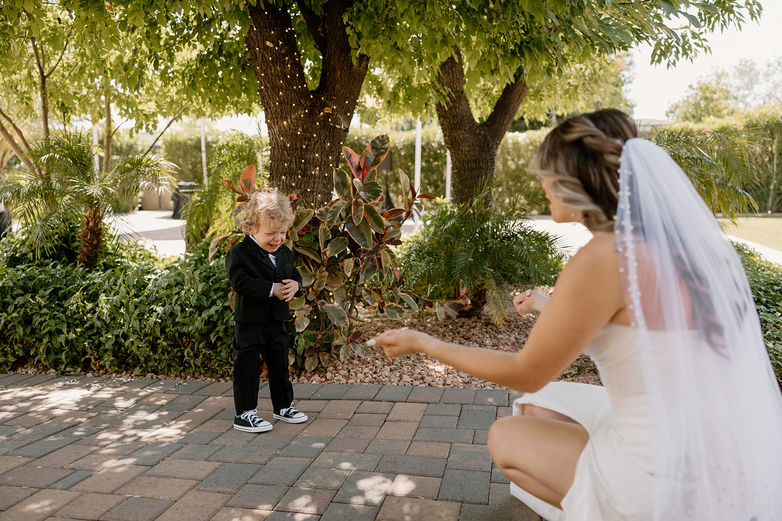 The bride kneels with open arms as a curly-haired ring bearer walks toward her in a tiny tux and Vans sneakers.