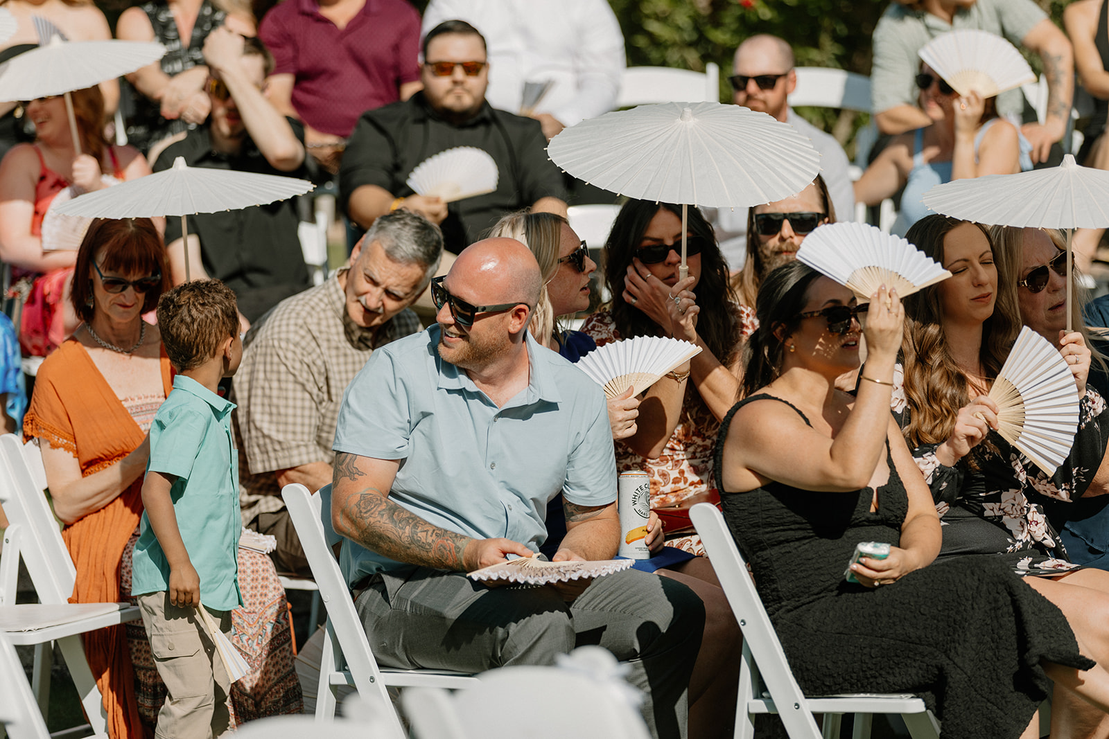 Guests shade themselves with paper parasols and fans, soaking in the ceremony under the desert sun.