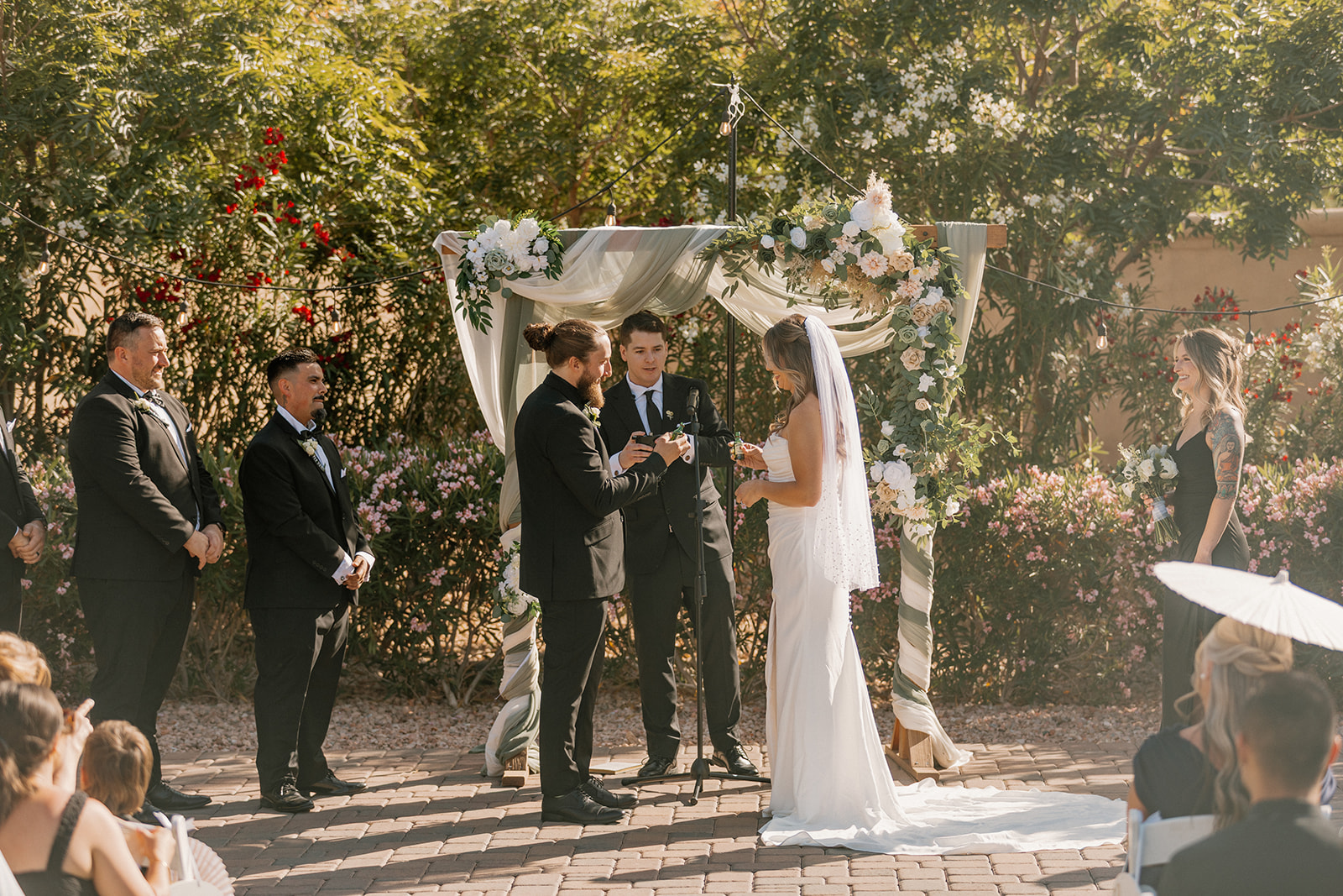 The couple stands beneath a flower-wrapped arch, holding hands and exchanging rings under the Arizona sun.