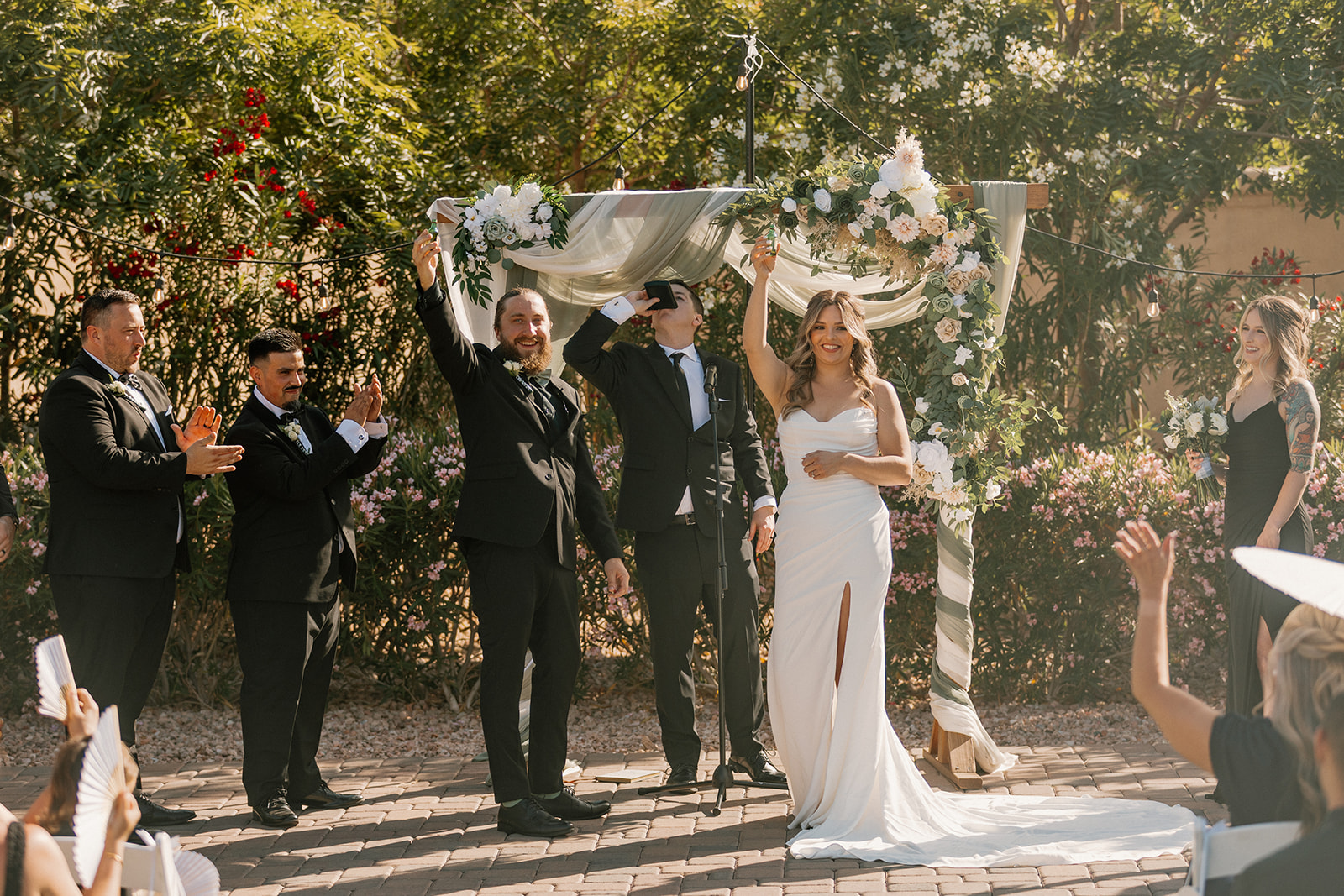 Just married! The couple raises their hands in celebration while guests cheer under a sun-drenched ceremony arch.