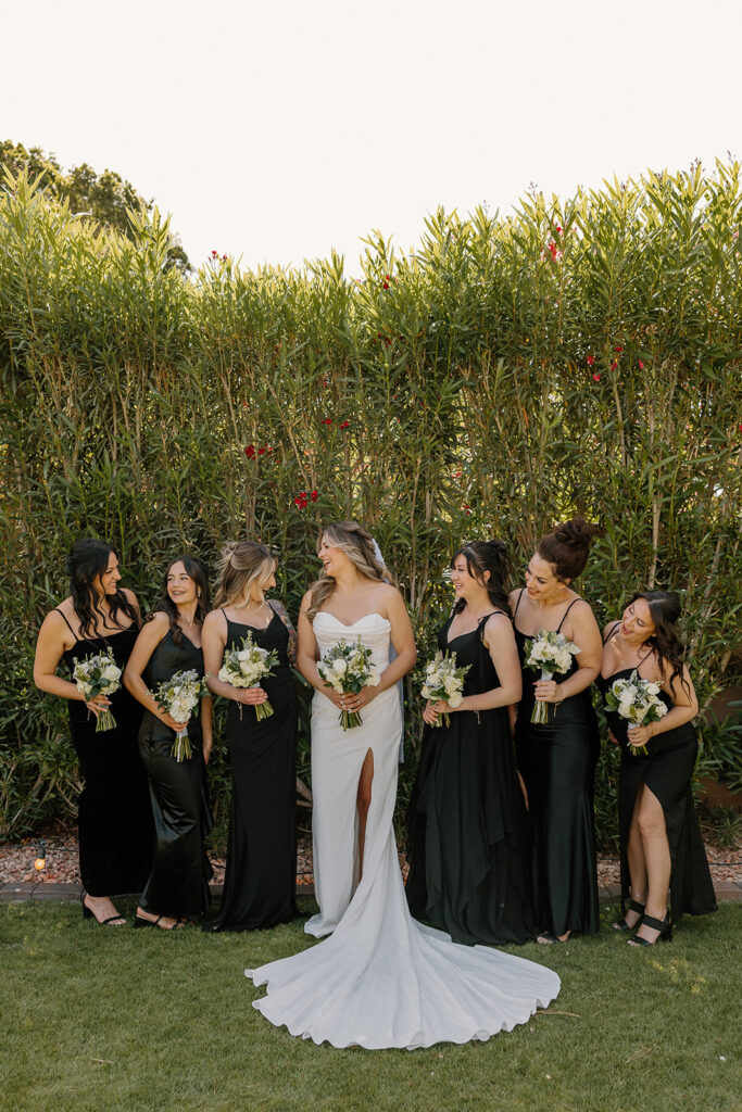 Bride and bridesmaids laugh together in matching black dresses, lined up under the afternoon sun.