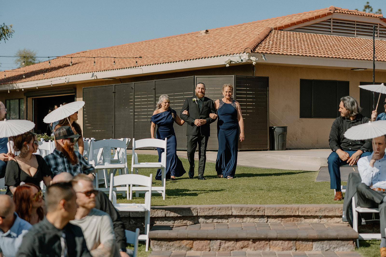 The groom’s mother and grandmother escort him down the aisle, all three smiling as they approach the crowd at an Arizona wedding venue.