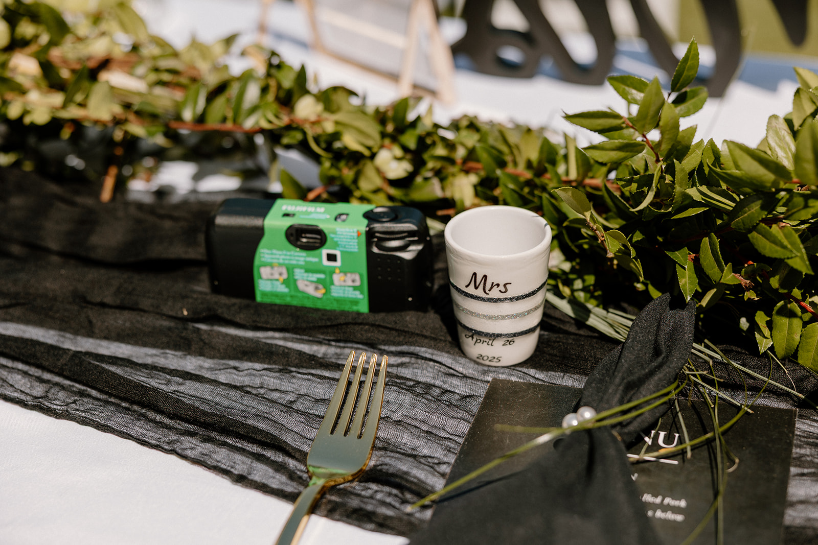 A disposable camera, personalized shot glass, and menu rest on the reception table surrounded by greenery at an Arizona wedding venue.