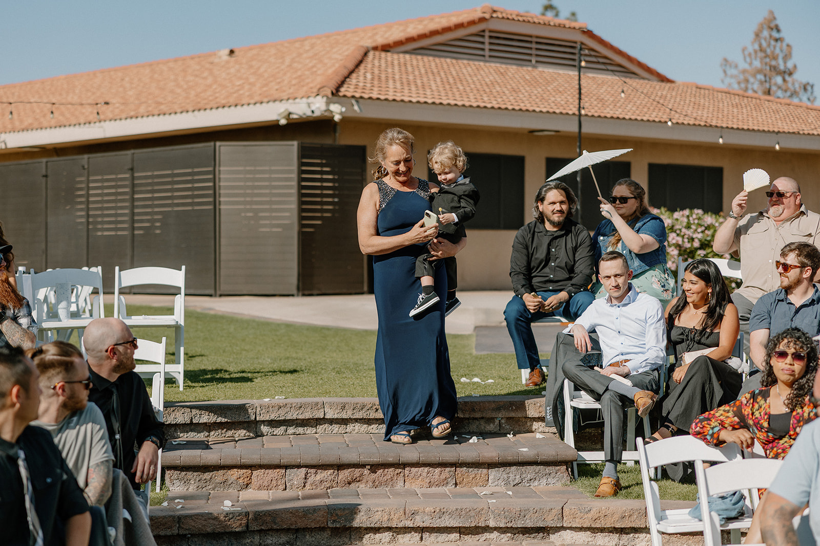 Ring bearer is carried by his grandmother as guests look on, waiting for the ceremony to begin.