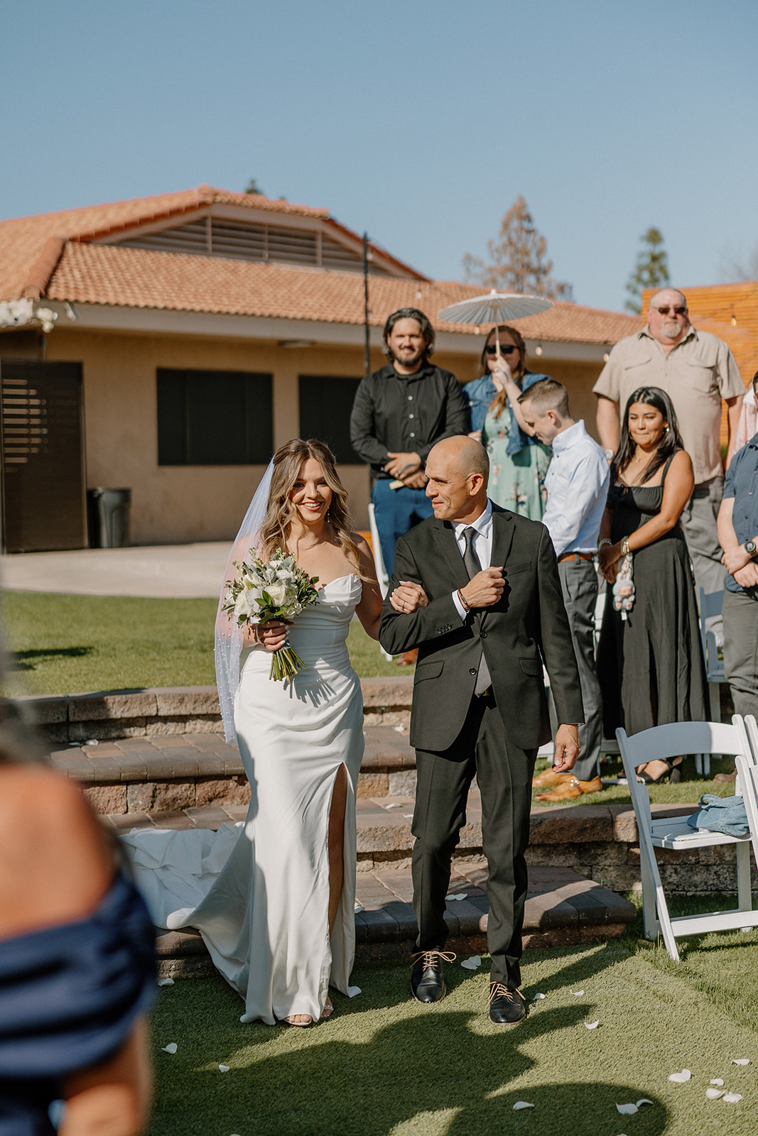 The bride walks down the aisle with her father, smiling at guests as they rise from their seats.