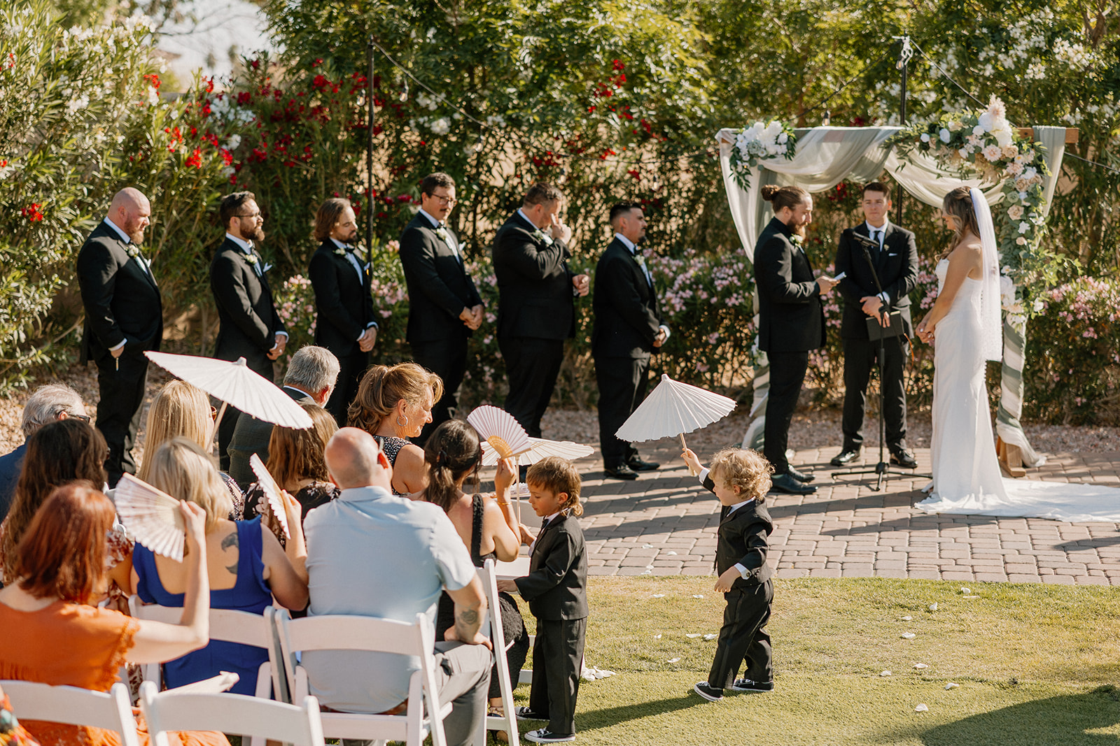The couple stands beneath a floral arch exchanging vows while guests fan themselves in the sun at their Arizona wedding venue.