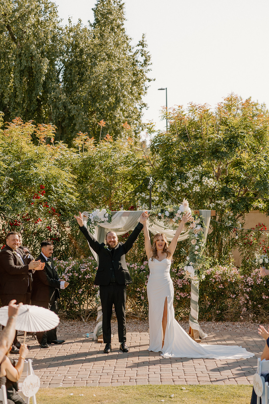 Hands in the air and big smiles—just married under a floral arch as guests cheer from the aisle.