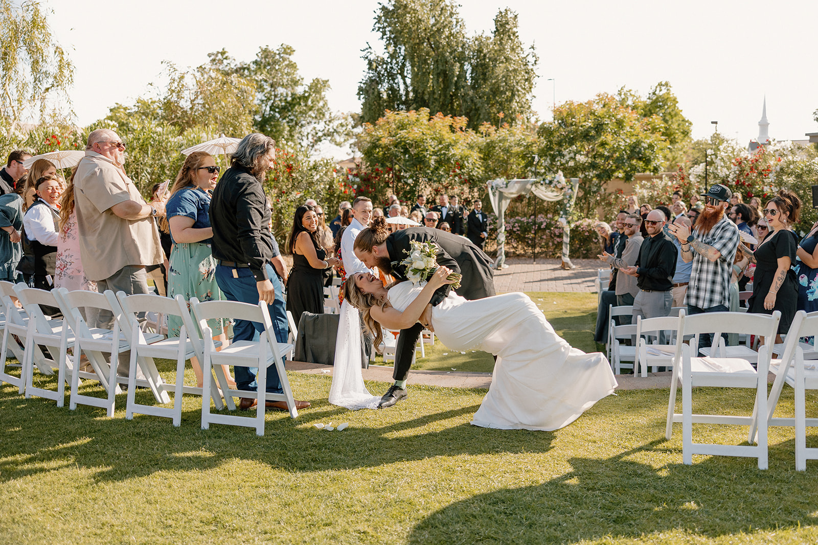 The groom dips the bride for a kiss during their joyful ceremony exit, surrounded by cheering guests at an Arizona wedding venue.