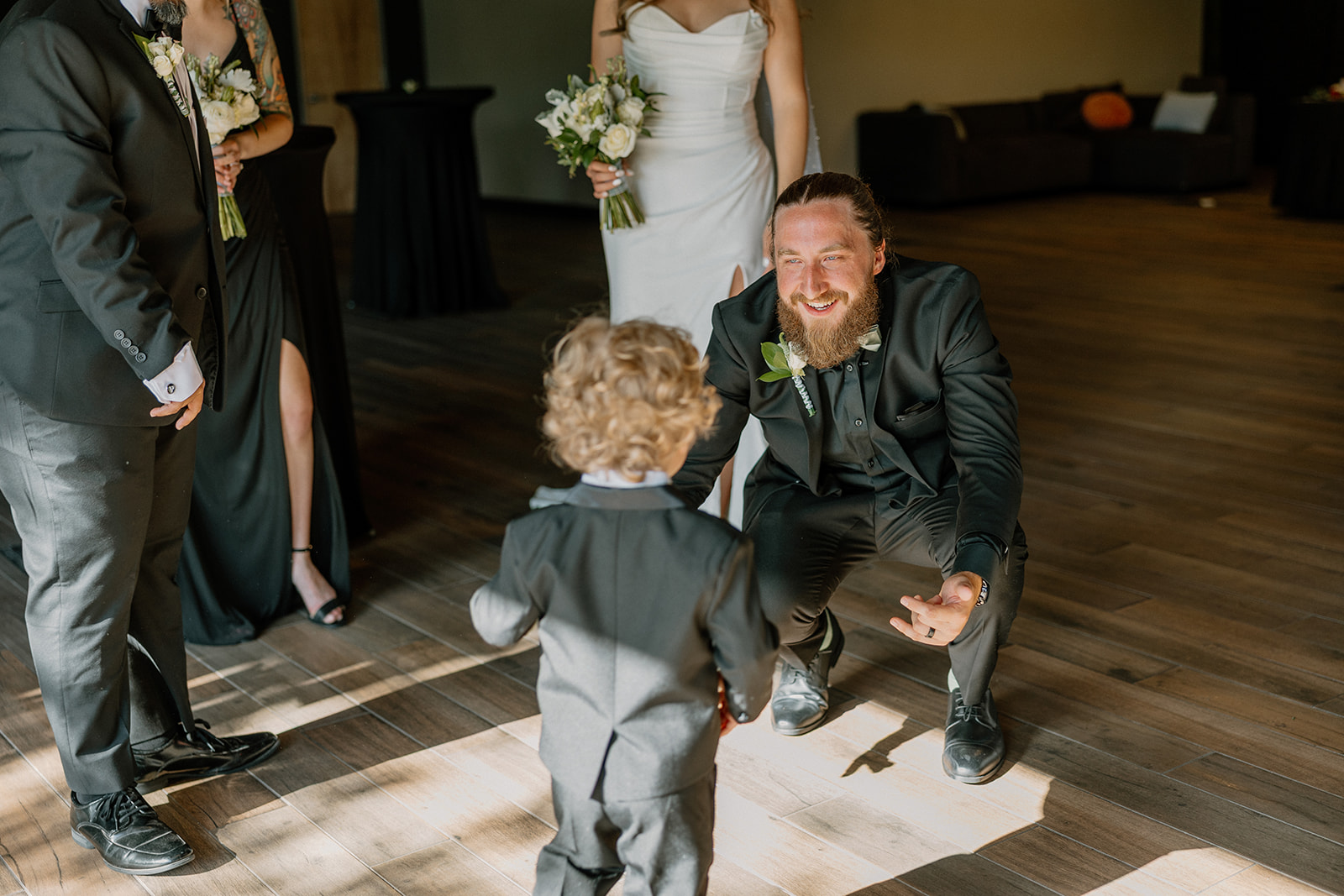 The groom kneels down with open arms to greet a curly-haired toddler in a miniature suit—pure sweetness.