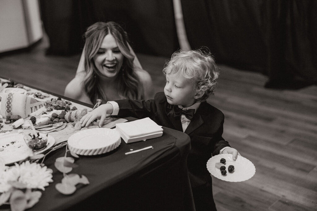 The bride crouches beside the dessert table, laughing as the ring bearer picks out snacks at their Arizona wedding venue.