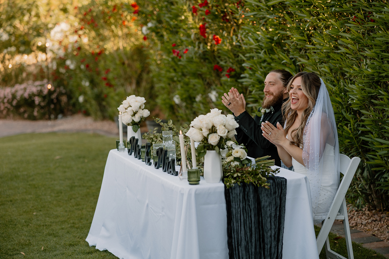 Bride and groom laugh and applaud at their sweetheart table, surrounded by white florals and candlelight.