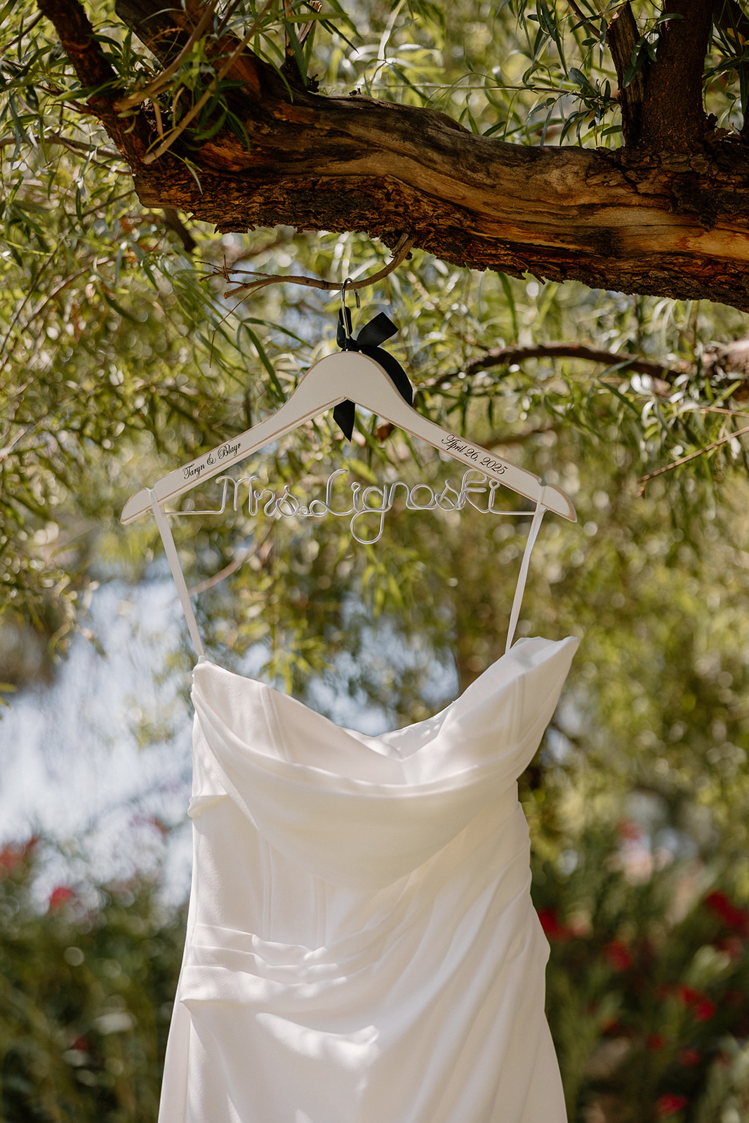 The bride’s dress hangs on a custom wire hanger from a tree branch, with light filtering through the leaves.