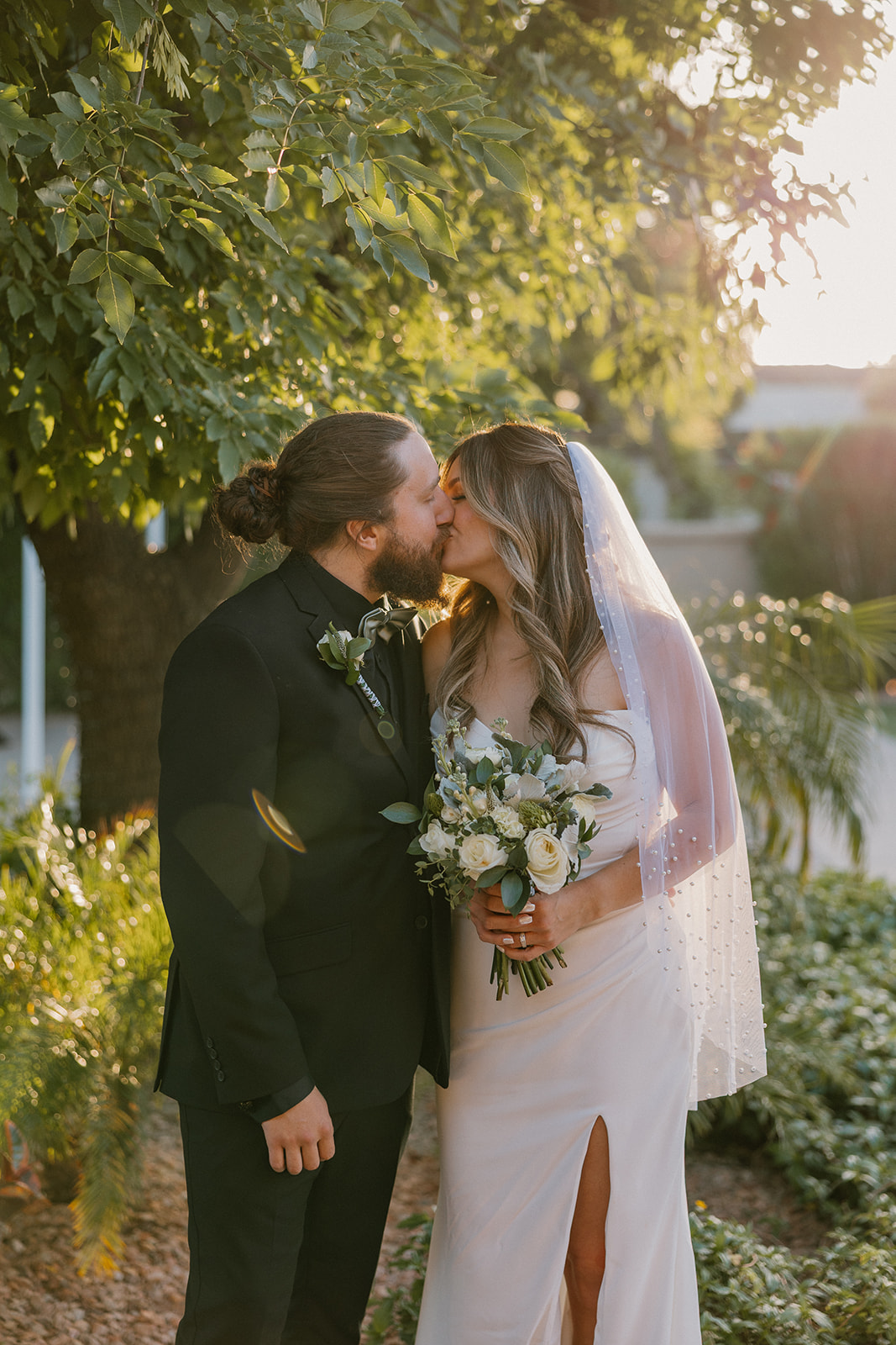 Newlyweds share a soft kiss surrounded by greenery as the sun glows behind them.