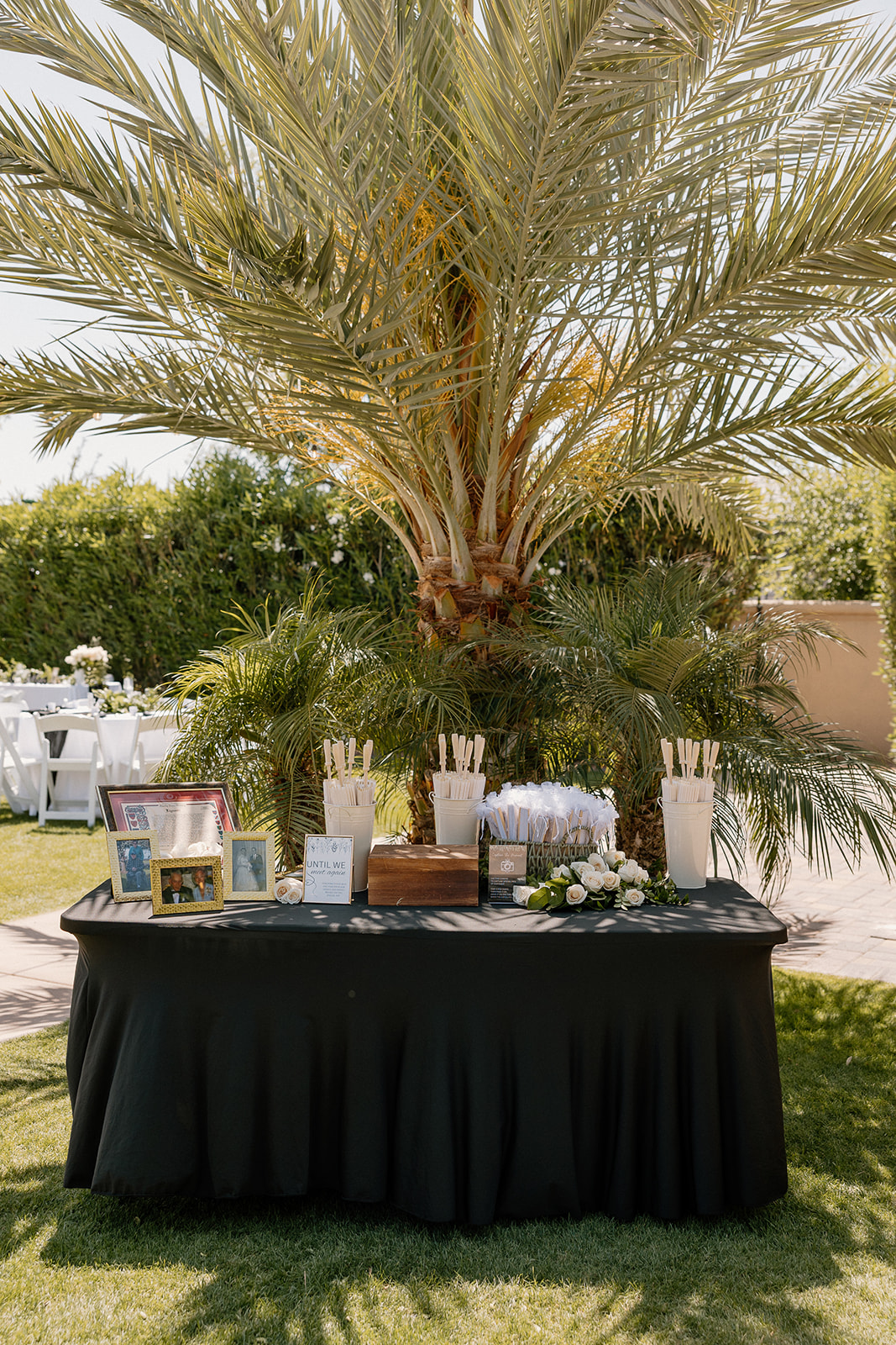 A tropical-inspired display table features wedding favors, parasols, and framed photos beneath a tall palm tree.