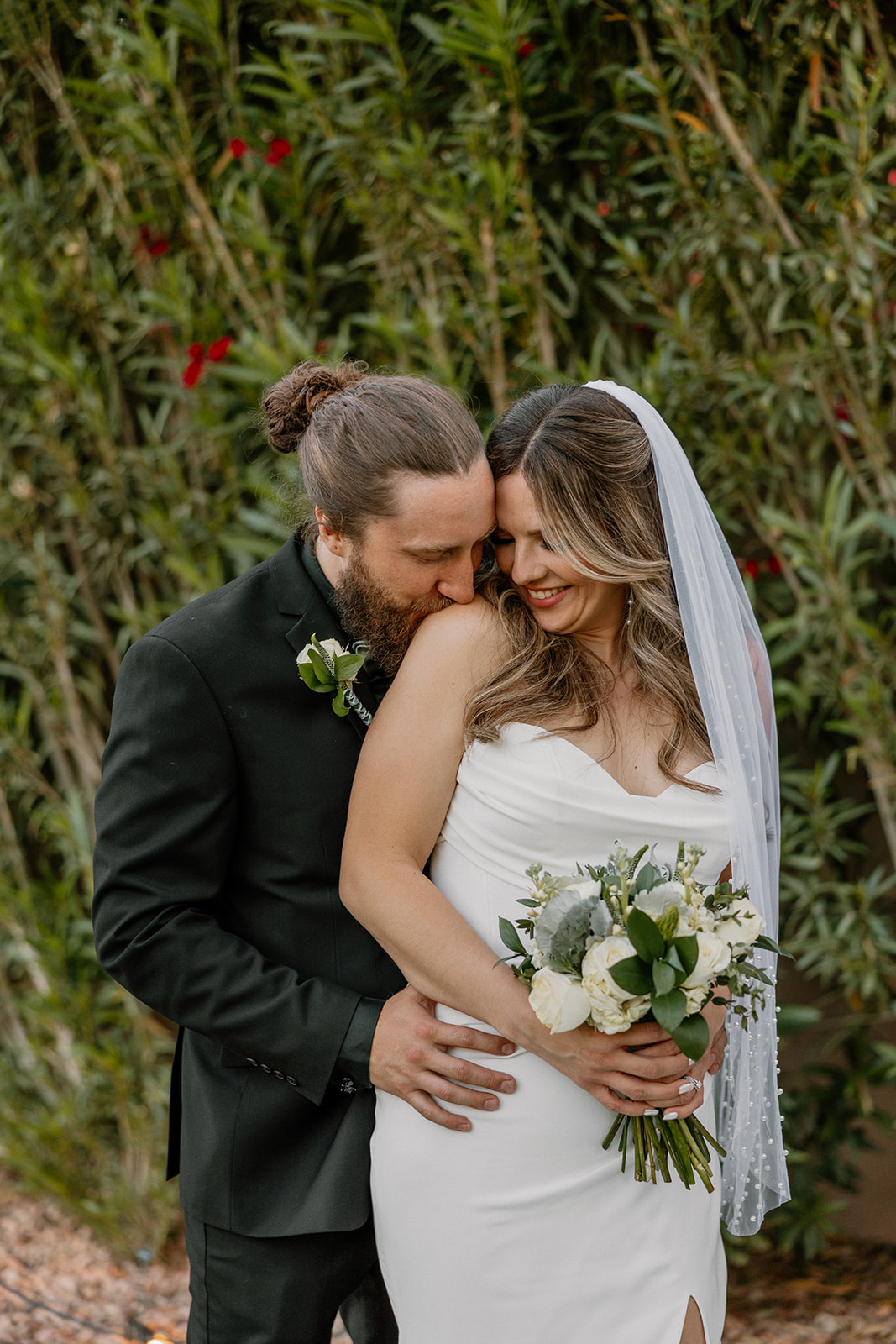 The groom wraps his arms around the bride and kisses her shoulder as they smile together in front of lush greenery.