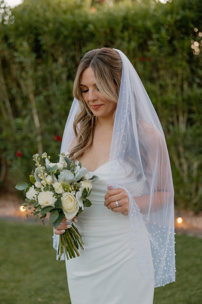 The bride admires her bouquet of white roses while standing alone in her flowing gown and pearl-trimmed veil.