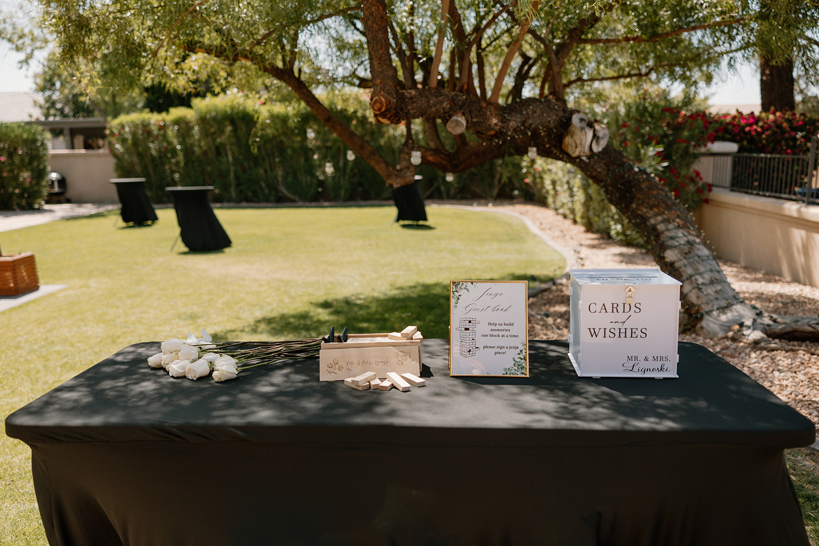 A welcome table is set beneath a shady tree, featuring a card box and custom Jenga guestbook at an Arizona wedding venue.
