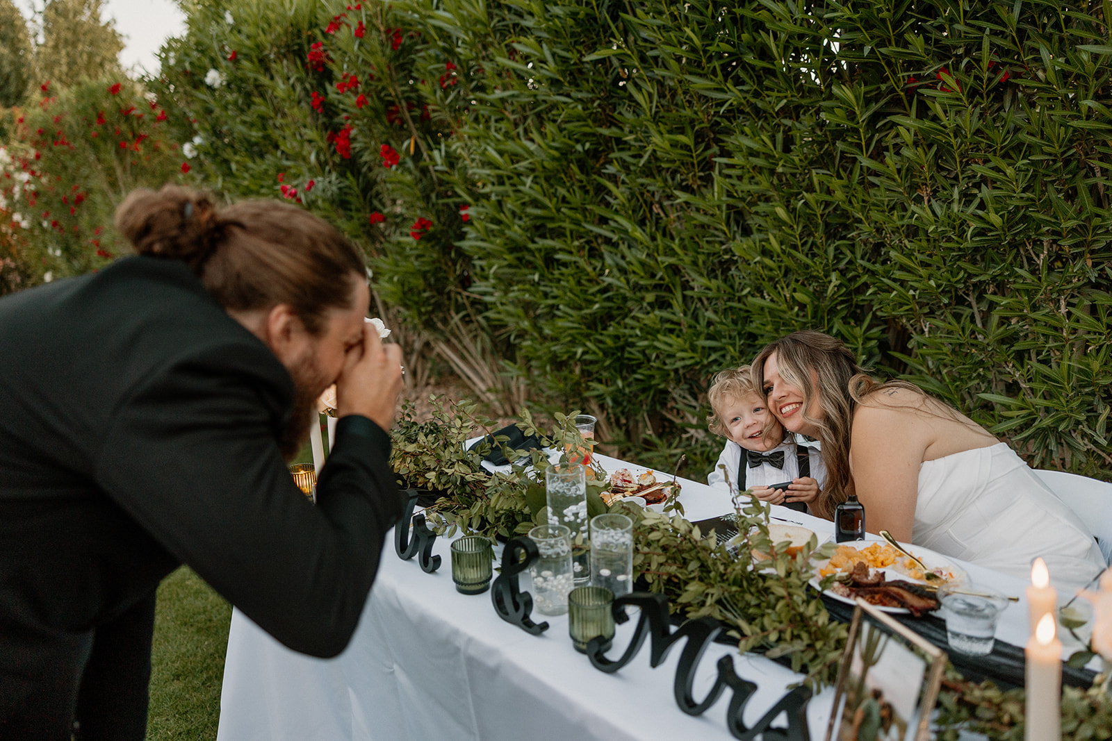 The groom takes a photo of the bride and their smiling son as they snuggle up at the sweetheart table.