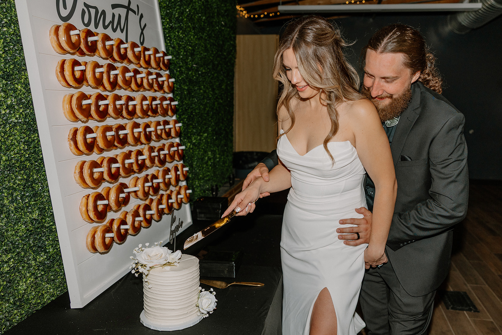The couple shares a playful moment while cutting their cake in front of a wall of donuts at their Arizona wedding venue.