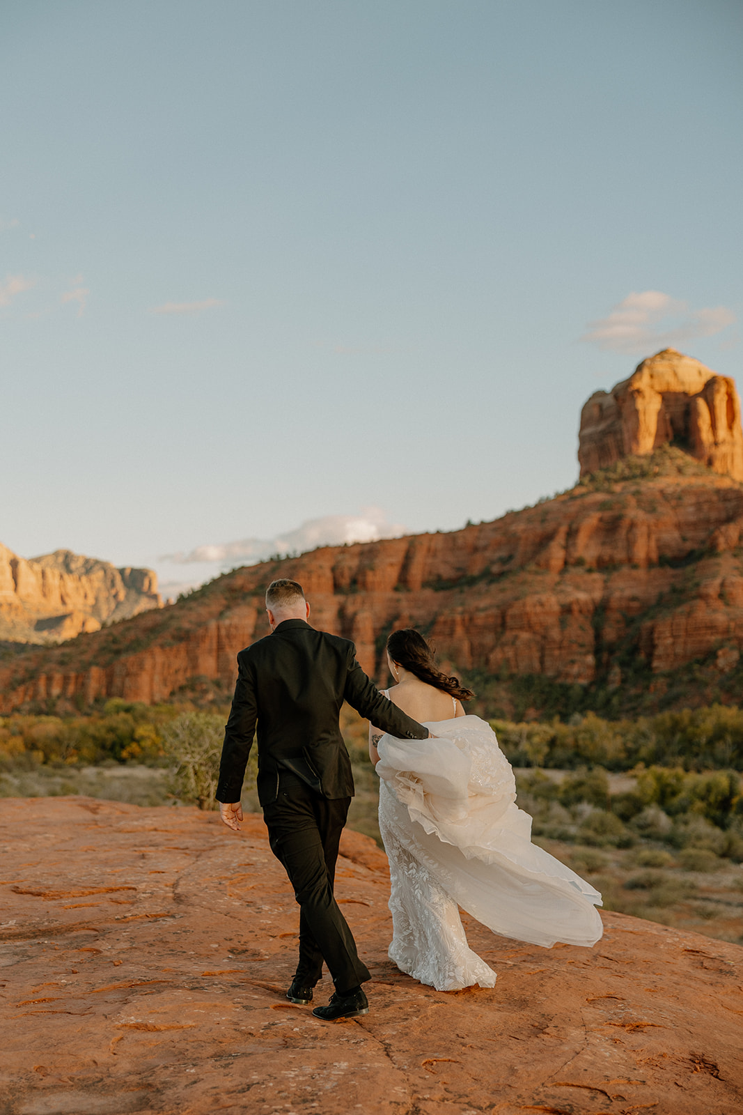 The couple walks hand-in-hand across red rock with her veil trailing behind them during their Sedona elopement.