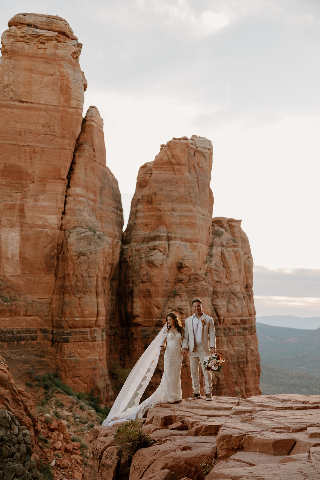 Bride and groom stand cliffside, her veil blowing in the wind as Cathedral Rock towers behind them—an epic Sedona elopement scene.