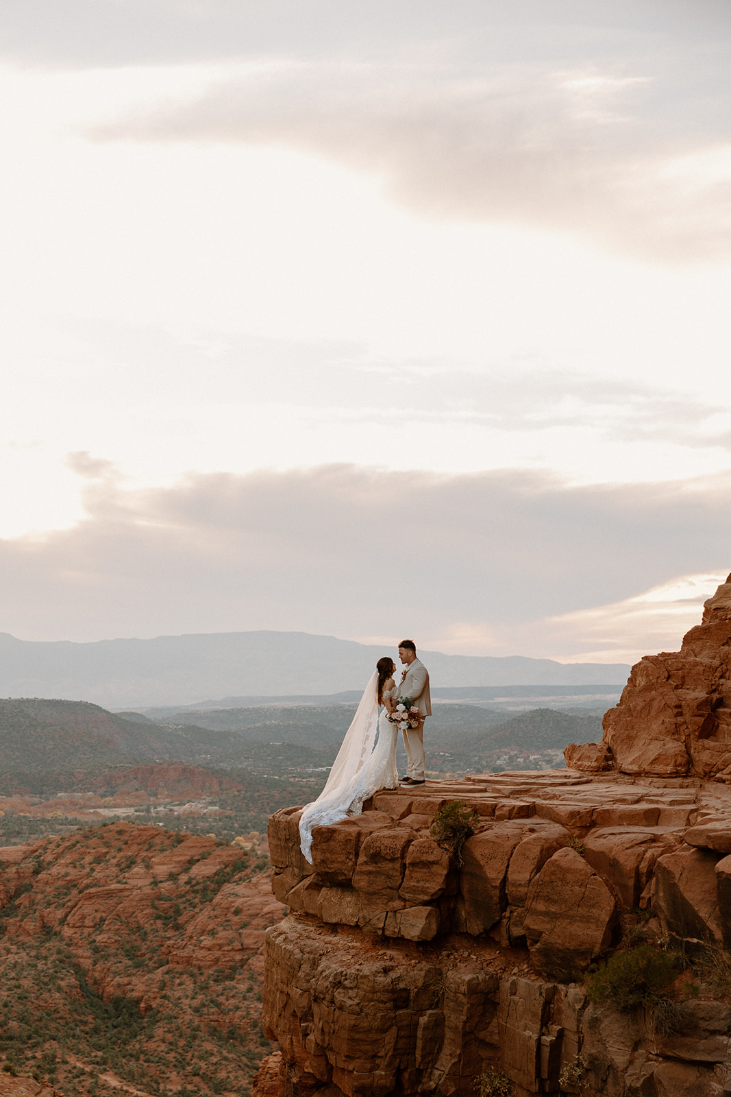 Perched on a ledge above the canyon, the couple shares a quiet kiss beneath a cloudy Sedona sky.